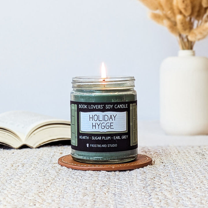 Candle labeled 'Holiday Hygge' on a coaster with a book and decorative elements in the background.