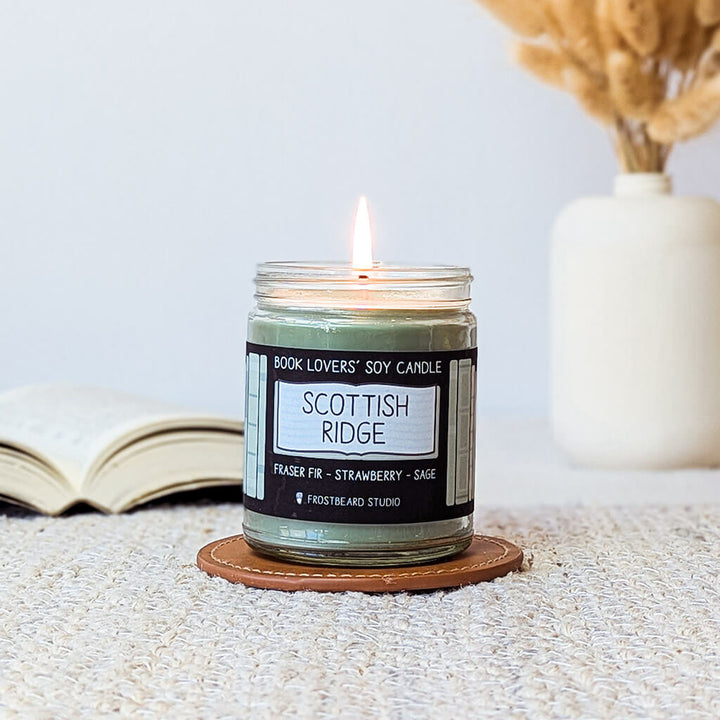 Candle labeled 'Scottish Ridge' on a coaster with an open book and decorative vase in the background.